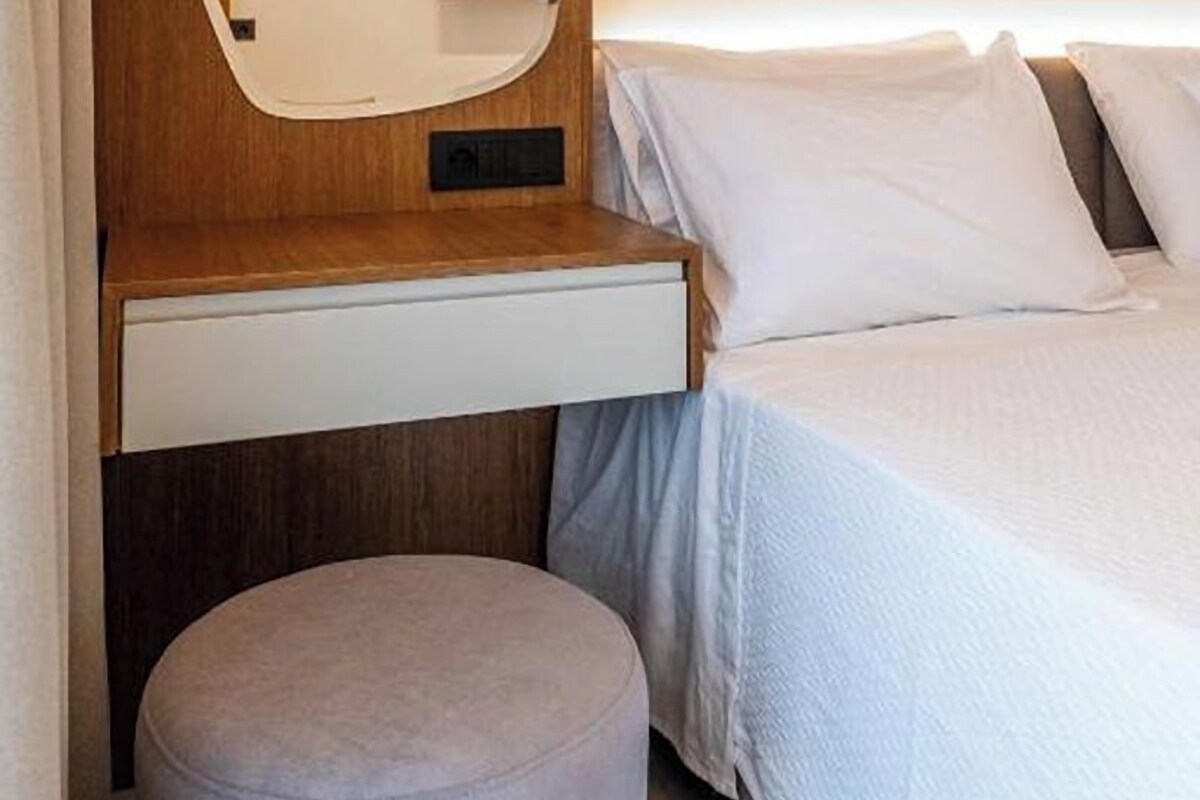 A modern bedroom with a wooden headboard, a bedside table, a round ottoman, and a bed with white linens.