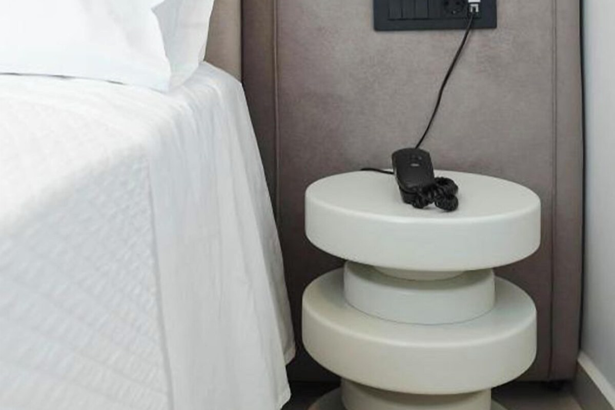 A bed with a white bedspread, a bedside table with a phone, and a power outlet on the wall.