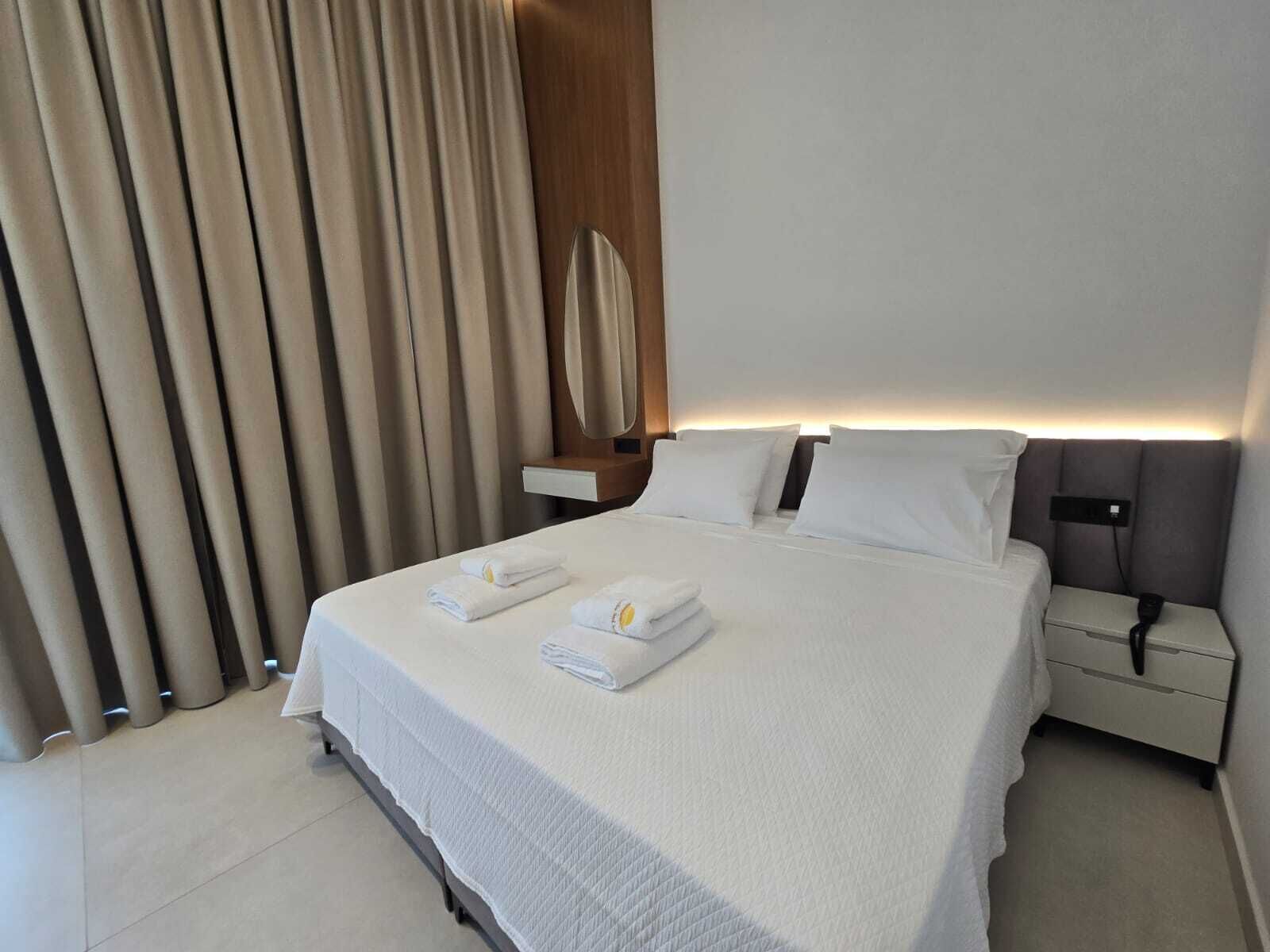 A hotel room with a neatly made bed, a bedside table with a lamp, and a large window with curtains.