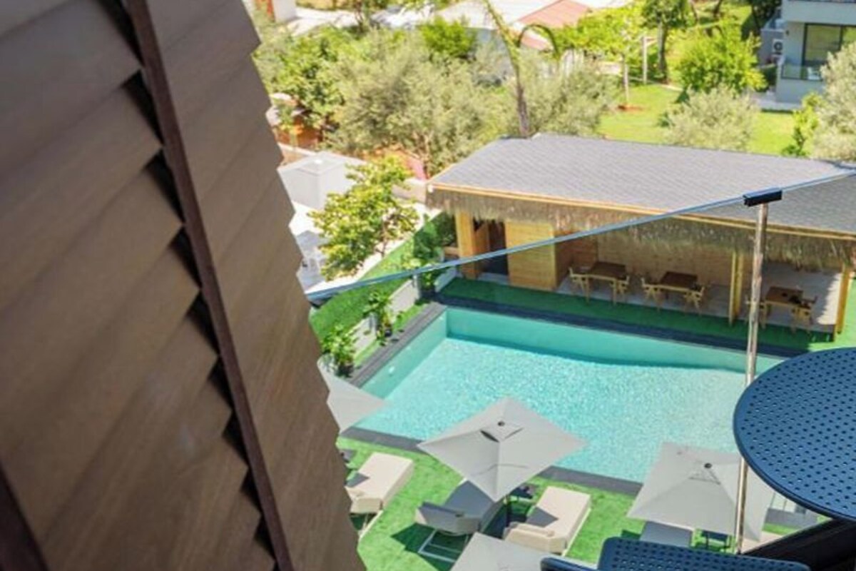 A view of a swimming pool with lounge chairs and umbrellas, surrounded by greenery and a thatched-roof structure.