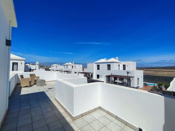 A rooftop terrace with white buildings, a clear blue sky, and a distant view of the landscape.