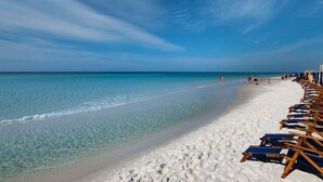 Une plage à l’eau turquoise et limpide, au sable blanc, bordée de rangées de chaises longues en bois.