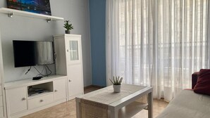 Una sala de estar con televisor, muebles blancos, una planta y cortinas de gasa.