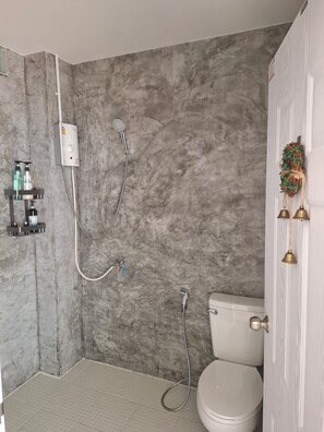 A bathroom with a grey marble wall, a white toilet, a shower with a handheld showerhead, and a shelf with toiletries. - A little Nap Hometel (Hat Yai)