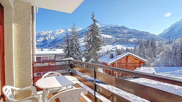 Ein Balkon mit Tisch und Stühlen, der auf eine verschneite Berglandschaft mit einer Holzhütte blickt.
