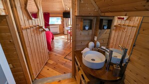 A wooden cabin interior with a bed, a small table, a sink, and a window.