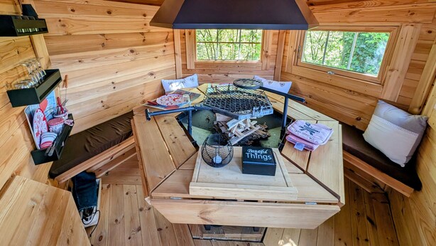 A wooden cabin interior with a central table, a grill, and a window overlooking greenery.