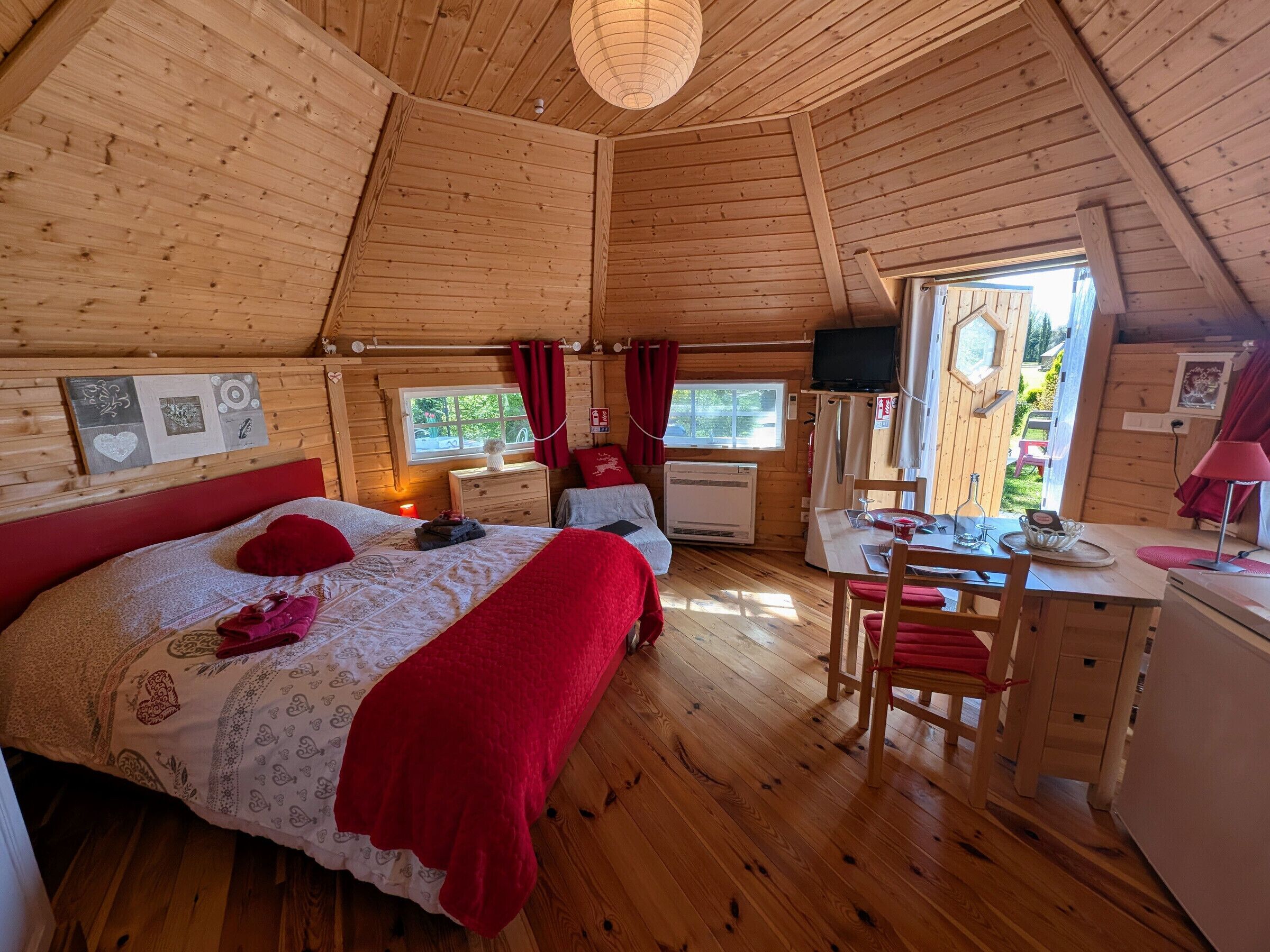 A cozy cabin with a wooden interior, a bed with red bedding, a desk, and a window overlooking a garden.
