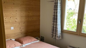 A single bed with a pink bedspread and pillows, a wooden wall, a window with curtains, and a radiator.