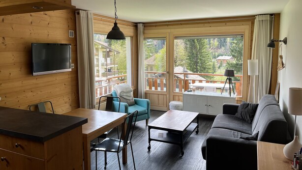 A room with a wooden interior, a sofa, a dining table with chairs, a TV mounted on the wall, and large windows with curtains.