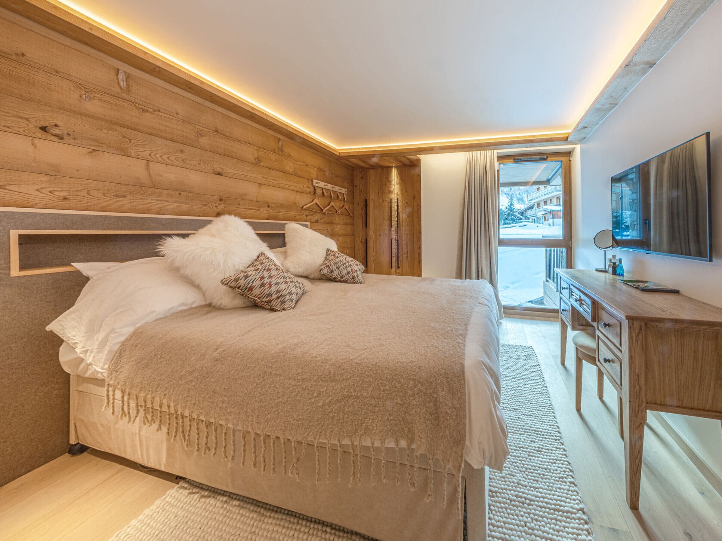 A bedroom with a wooden headboard, a bed with white and patterned pillows, a wooden desk, and a large window offering a view of a snowy landscape.