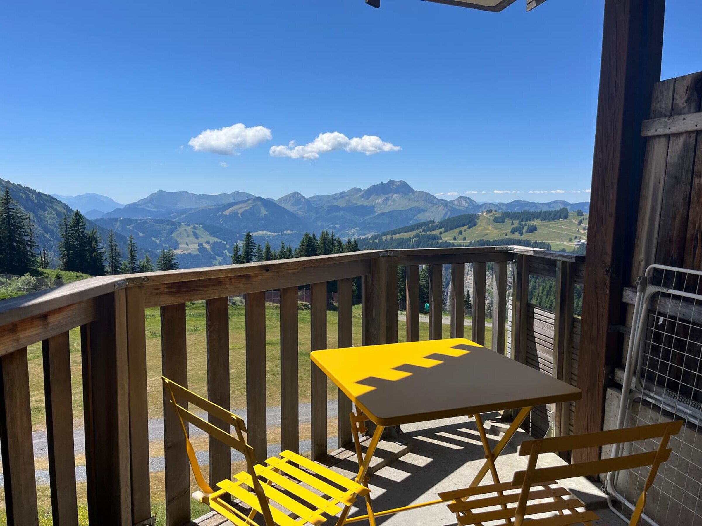 A wooden deck with a yellow table and chairs, overlooking a mountainous landscape.