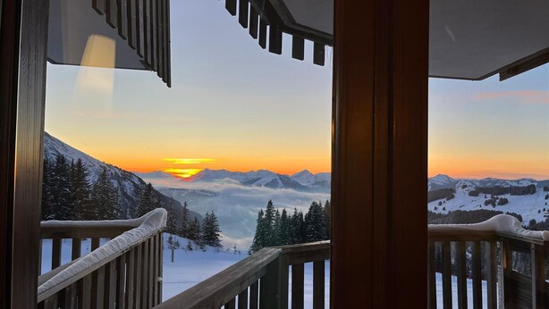 Een houten trap leidt naar een balkon met uitzicht op een besneeuwd berglandschap in de zonsondergang.