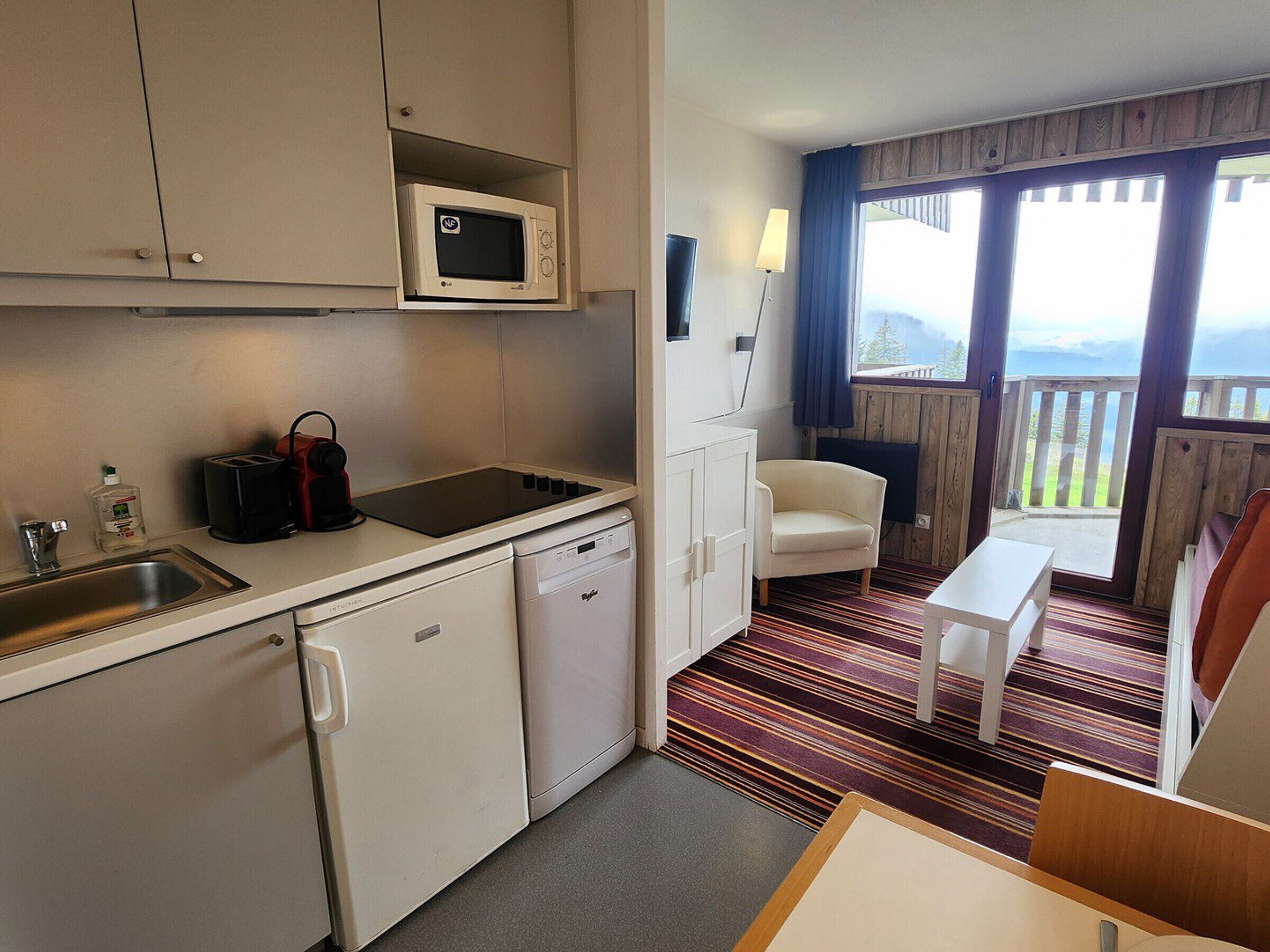 Eine kompakte Küche mit Mikrowelle, Toaster, Spüle und Geschirrspüler. Es gibt einen kleinen Essbereich mit Tisch und Stuhl, und ein Fenster mit Blick ins Freie.