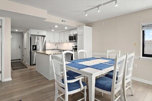 Une cuisine moderne avec des meubles blancs, des appareils en acier inoxydable et un coin repas agrémenté d’un chemin de table bleu et de chaises blanches.