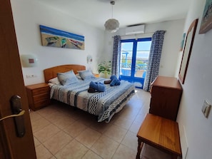 Un dormitorio con cama, mesitas de noche, cómoda, un cuadro y vistas a través de una ventana.