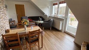 Un coin repas avec une table et des chaises en bois, une petite table d’appoint sur laquelle est posé un vase de fleurs, et une fenêtre avec des stores.