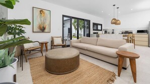 Un salon moderne avec un canapé beige, une table basse ronde et des chaises en bois. Un grand tableau abstrait orne le mur, et on aperçoit un coin cuisine au fond.