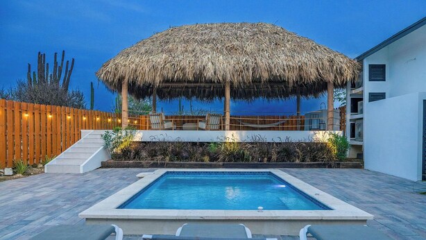 Une piscine avec des chaises longues, une structure dotée d’un toit de chaume et une clôture en bois.