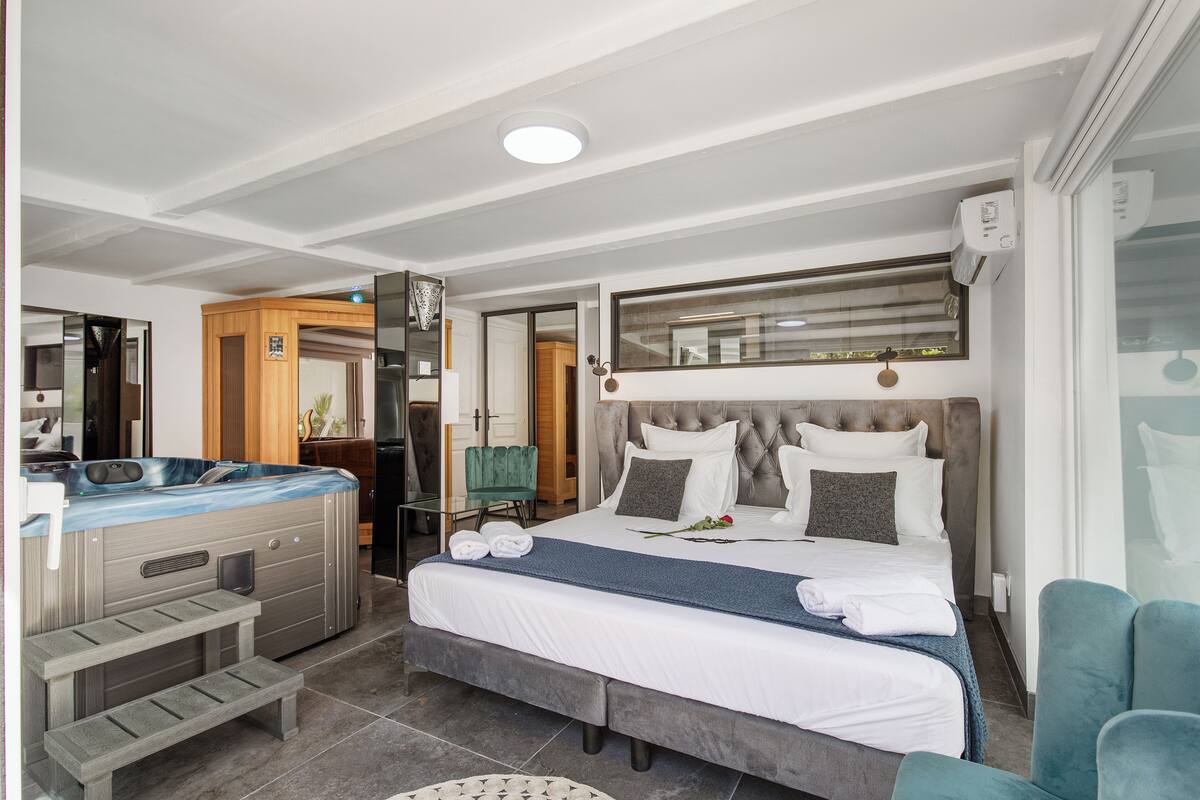 A modern hotel room with a large bed, a hot tub, a seating area, and a mirror above the bed.