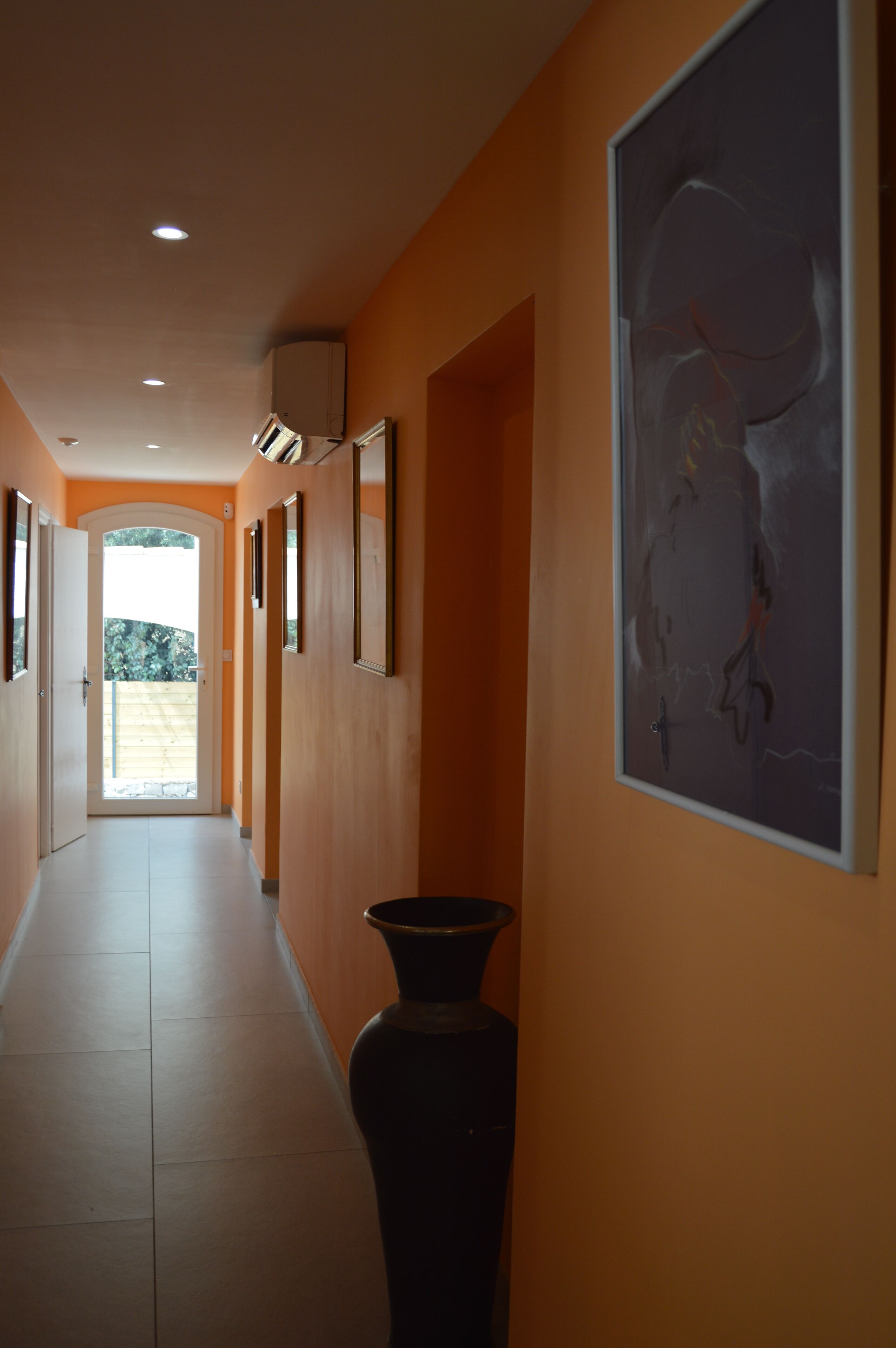 A hallway with a long, narrow layout, featuring a large vase and framed artwork on the walls.