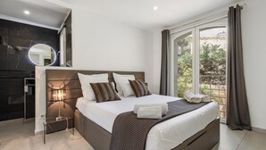 A modern bedroom with a large bed, a bedside table, a mirror, and a view of the outdoors. - VILLA RASOA (Agde)