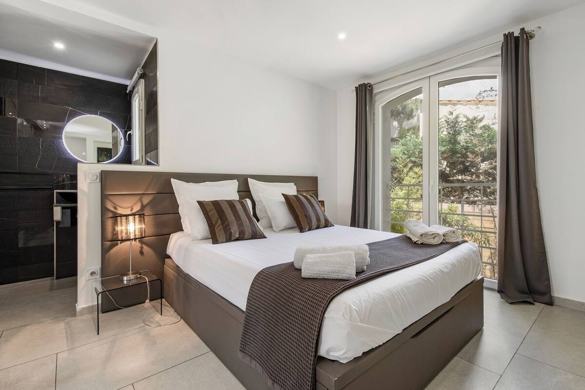 A modern bedroom with a large bed, a bedside table, a mirror, and a view of the outdoors.