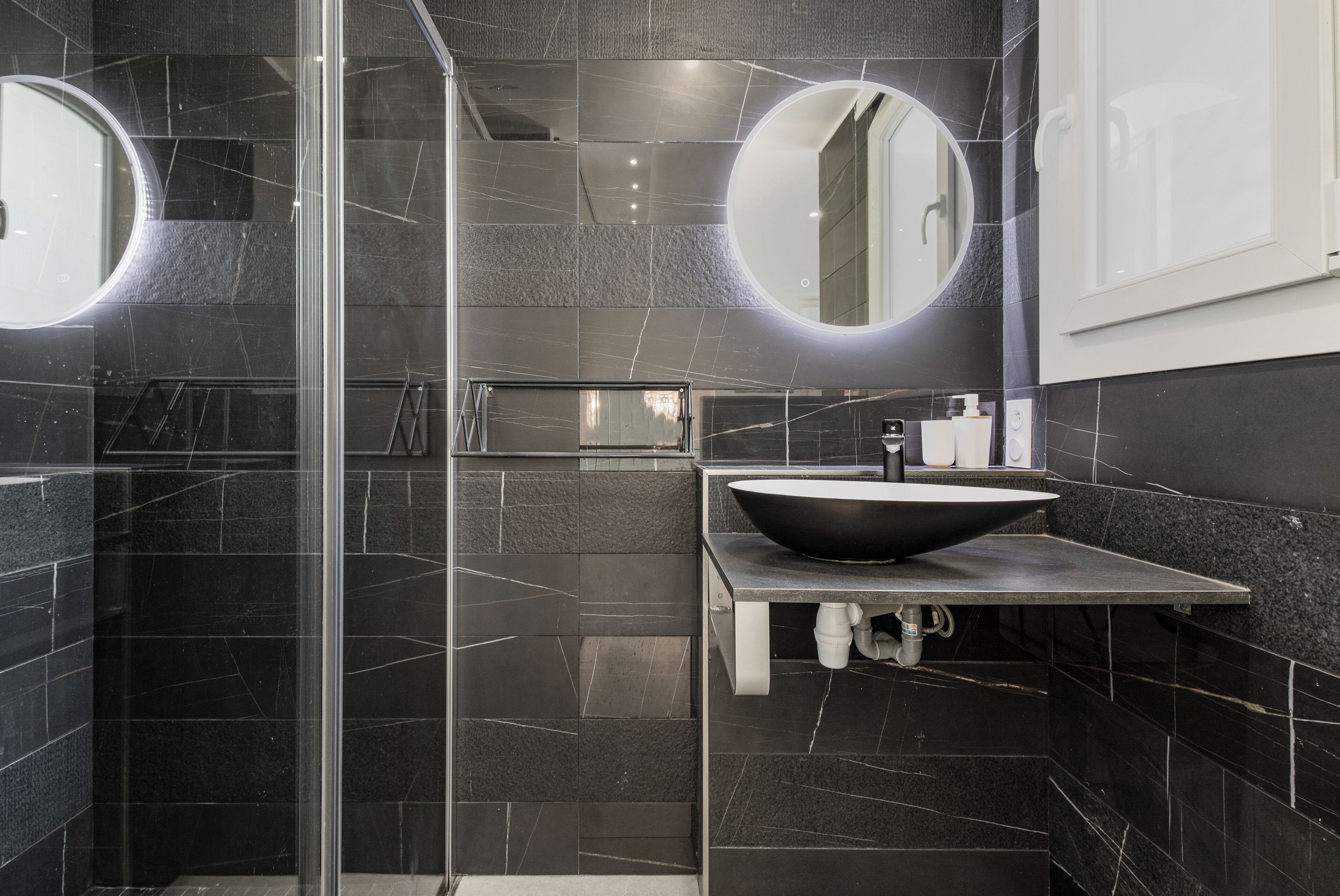 Un baño moderno con baldosas oscuras, un lavamanos grande y dos espejos redondos.