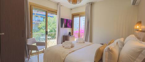A hotel room with a large bed, a chair, a TV, and a balcony with a view of buildings and trees.