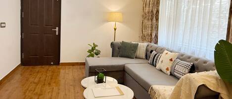 Una sala de estar moderna con un sofá gris, una mesita redonda pequeña, una lámpara y un ventanal con cortinas translúcidas.