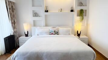A modern hotel room with a large bed, white bedding, two bedside lamps, a radiator, a small plant, and decorative items on shelves.