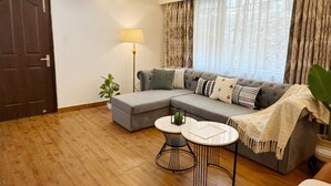 Een woonkamer met een hoekbank, een ronde salontafel, een vloerlamp en een groot raam met gordijnen.