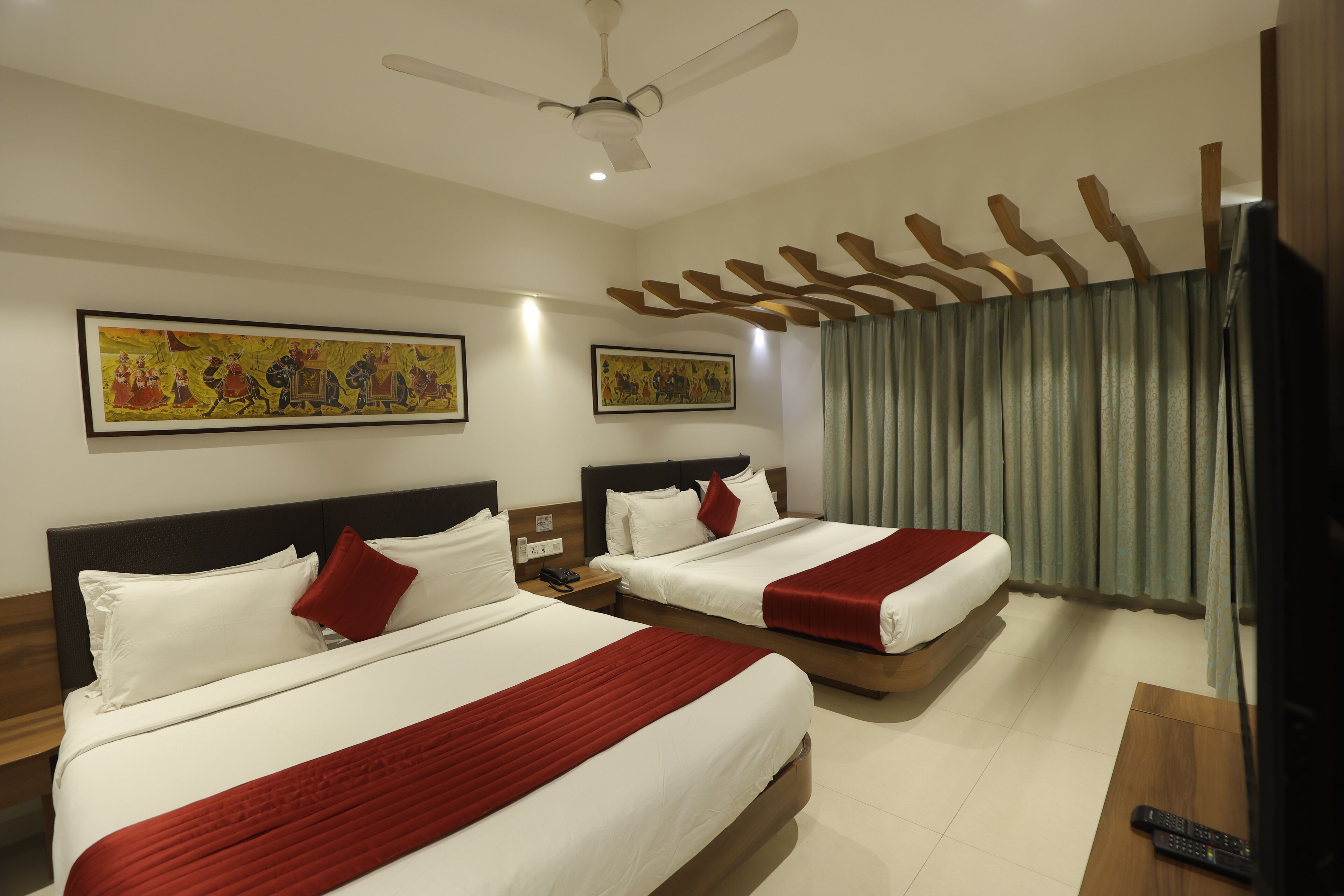A hotel room with two beds, a ceiling fan, artwork on the walls, and a television.