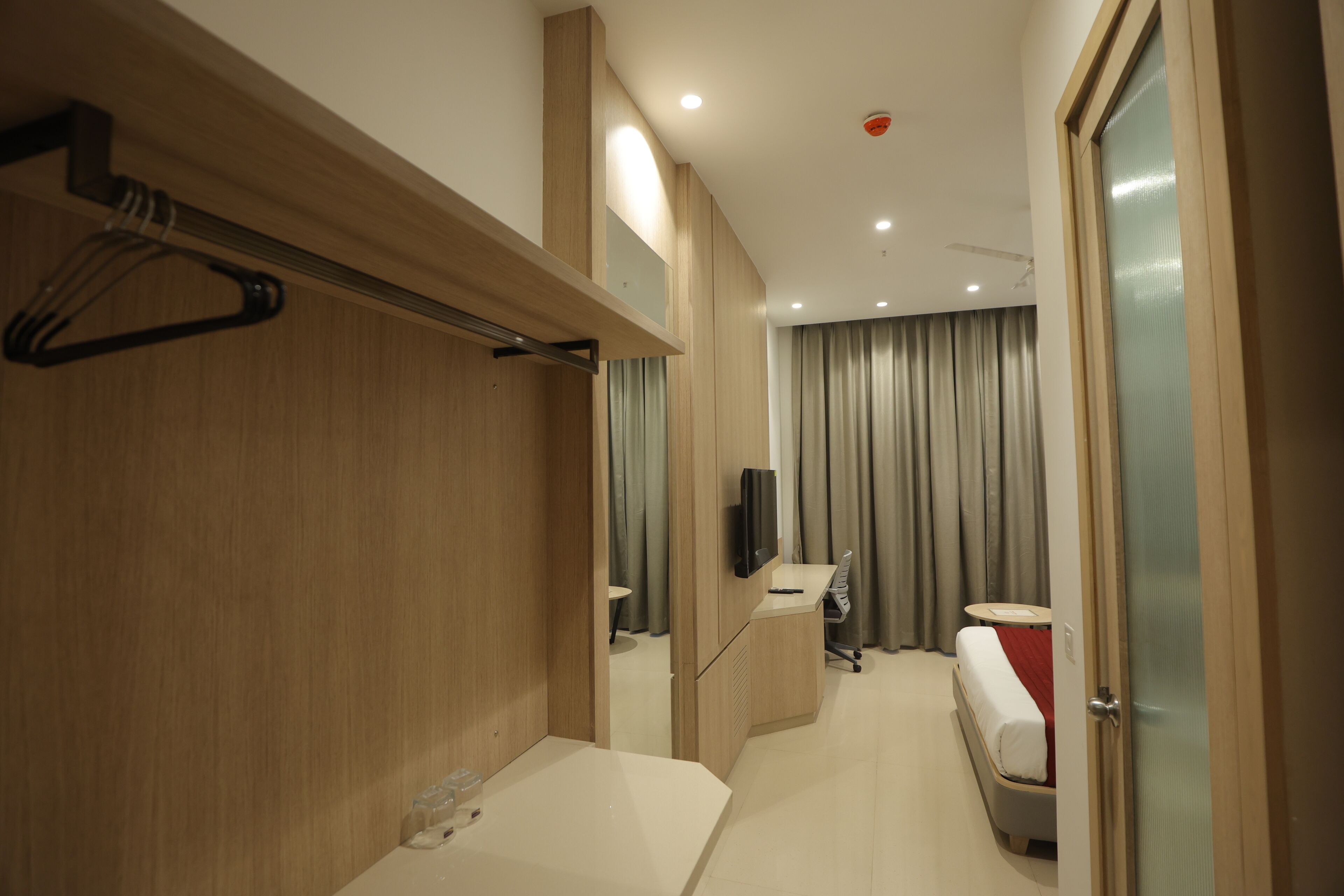 Een moderne hotelkamer met een houten opbergmeubel, een bed met een rode deken, een bureau met een stoel, en een televisie die aan de muur is bevestigd.