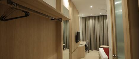 Een moderne hotelkamer met een houten opbergmeubel, een bed met een rode deken, een bureau met een stoel, en een televisie die aan de muur is bevestigd.