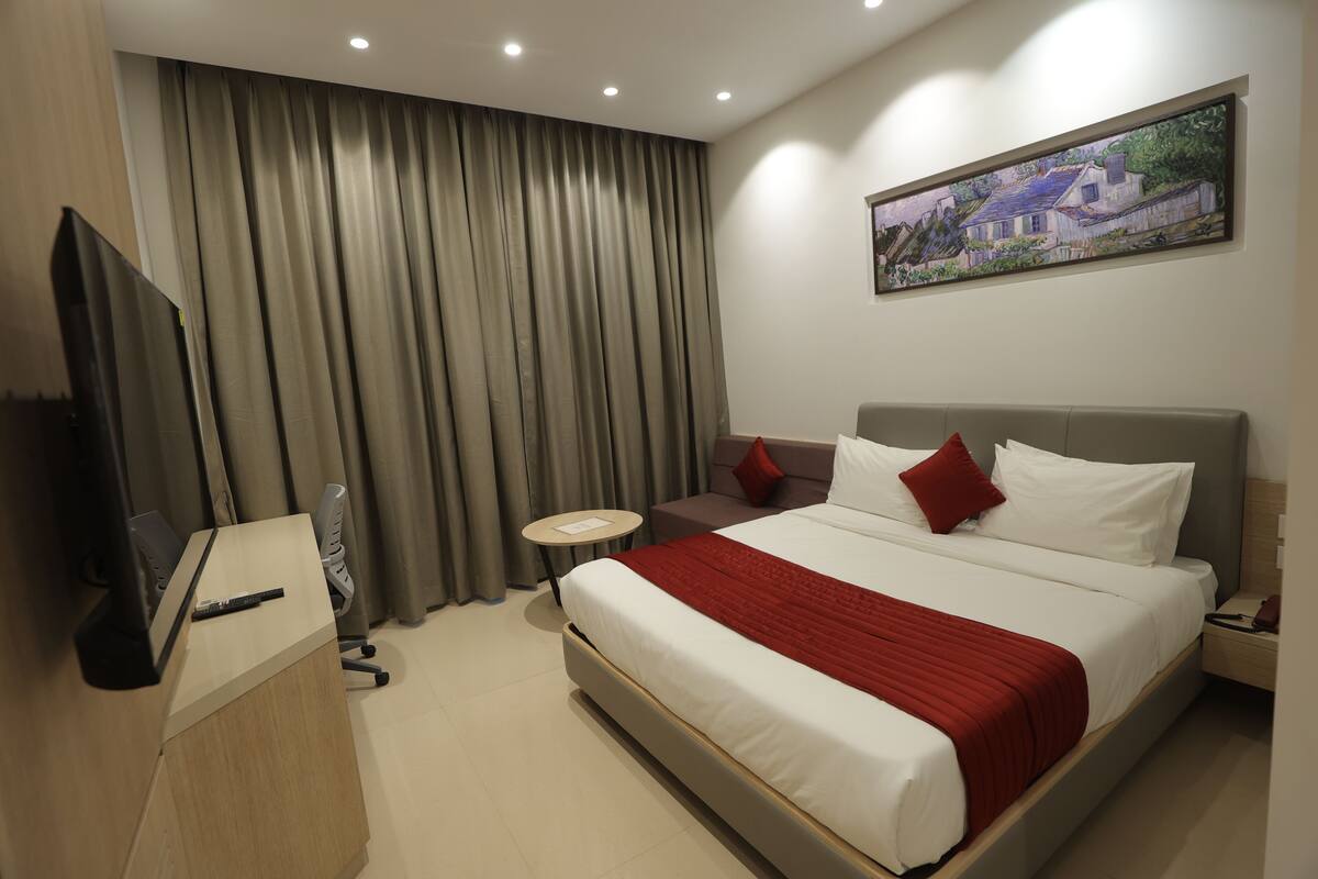 A hotel room with a large bed, a desk, a television, a small round table, and a painting on the wall.