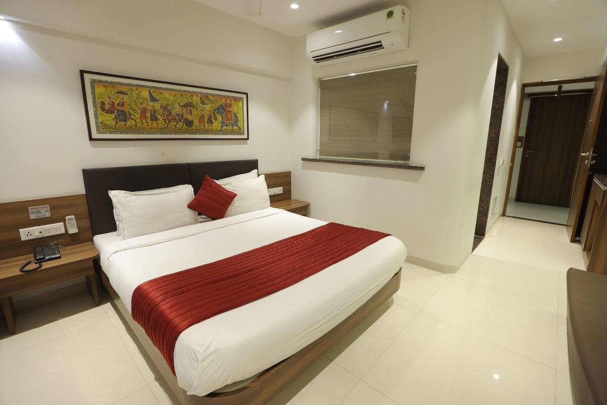 A hotel room with a large bed, a nightstand with a phone, a window with blinds, and a wall-mounted air conditioner.