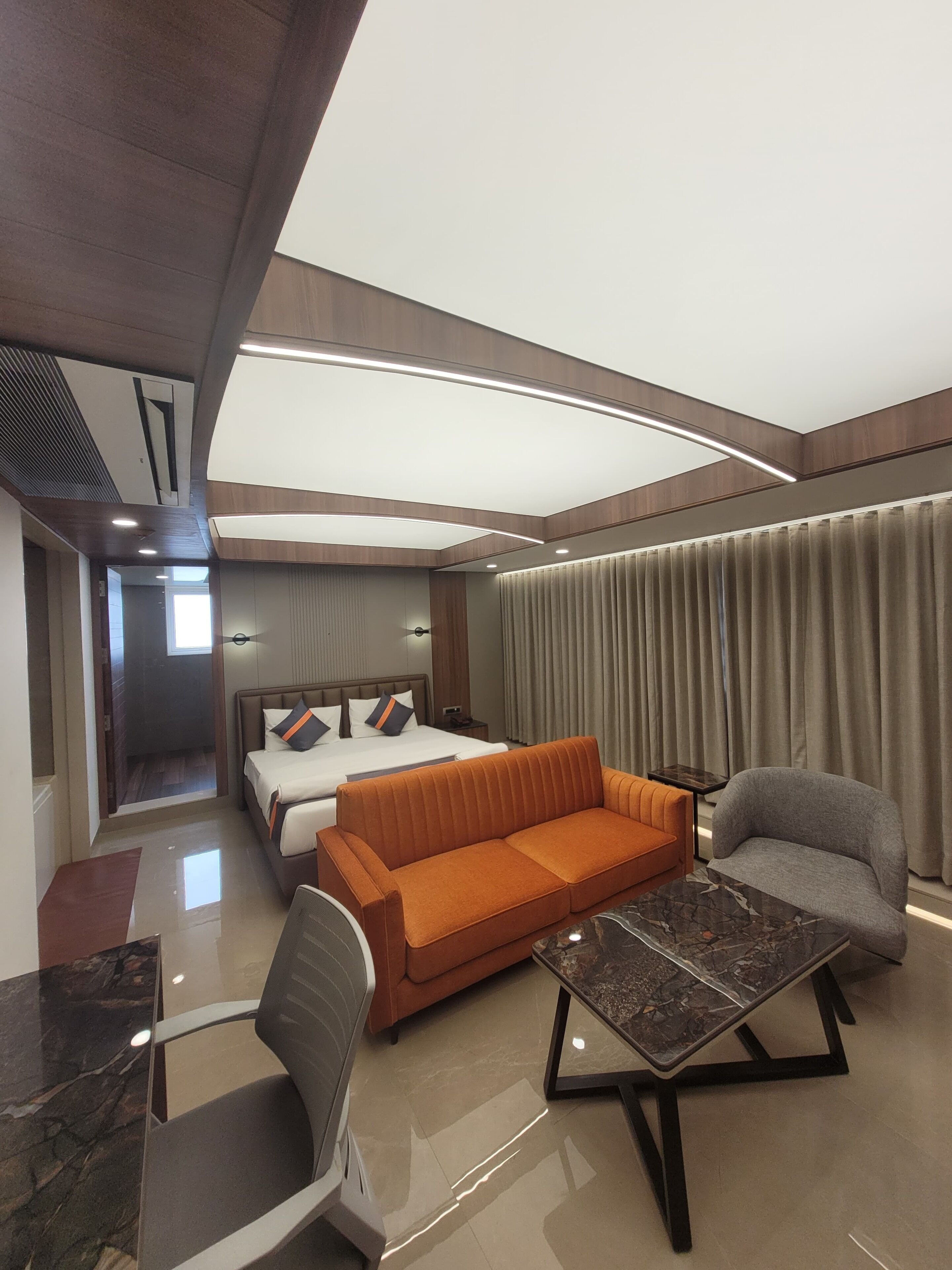 Une chambre d’hôtel moderne avec un lit, un canapé orange, un fauteuil gris et une table basse en marbre.