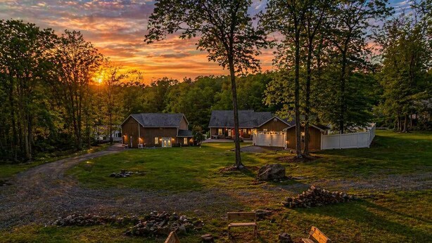 A rustic lodge with a fire pit and benches, surrounded by trees and a sunset view.