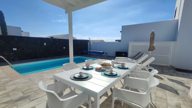 A covered outdoor dining area with a pool, white furniture, and a clear sky. - Villa Anny, heated pool, ideal for families! (PLAYA BLANCA)