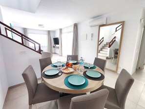 Un coin repas avec une table ronde dressée pour quatre personnes, des chaises, une bouteille d’eau et un saladier de fruits.