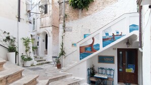A narrow street with steps leading to a building with a wooden door and a sign that reads "Vino". There are potted plants and a table with chairs outside. - Casa Vacanza Opuntia (Sperlonga)