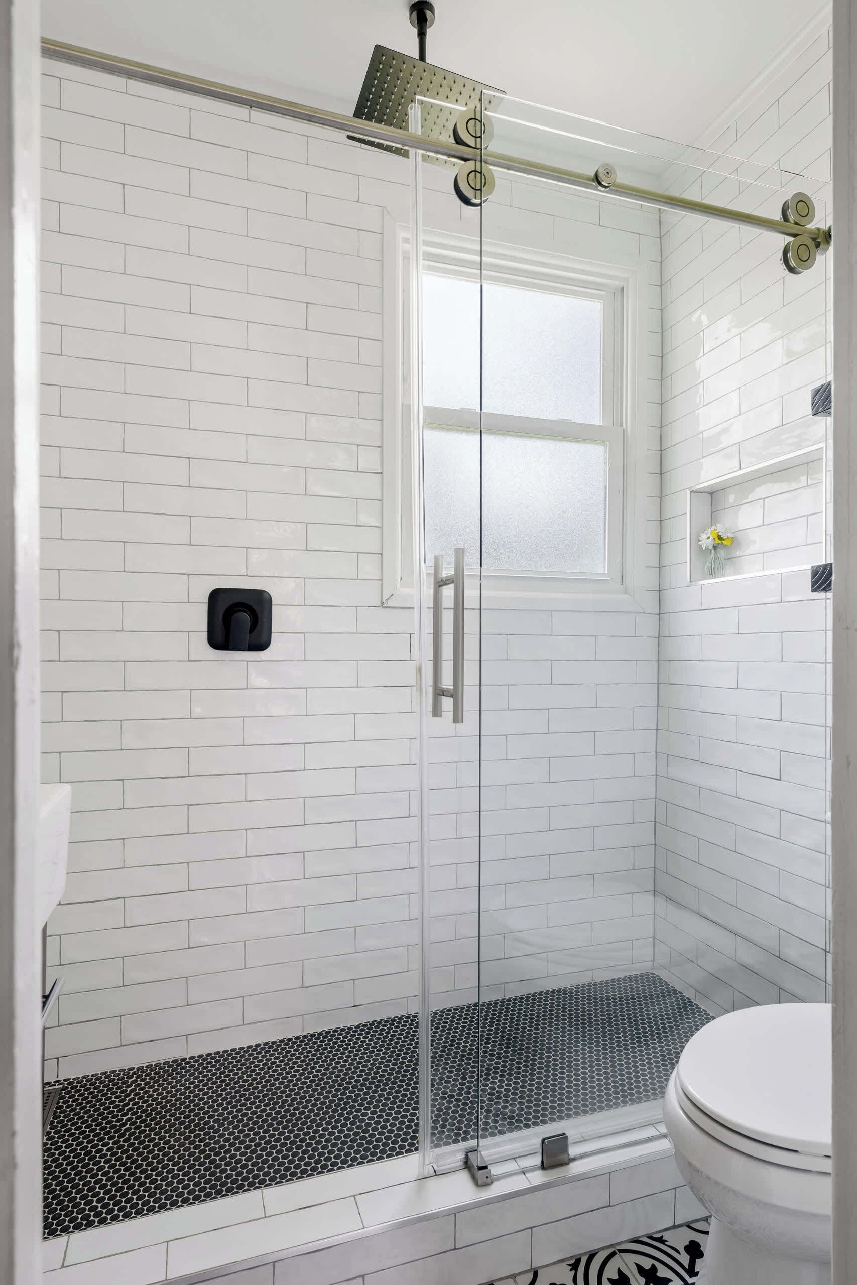 Ein Badezimmer mit weißen Fliesen, einer Glasdusche, einer weißen Toilette und einem schwarz-weißen Bodenbelag.
