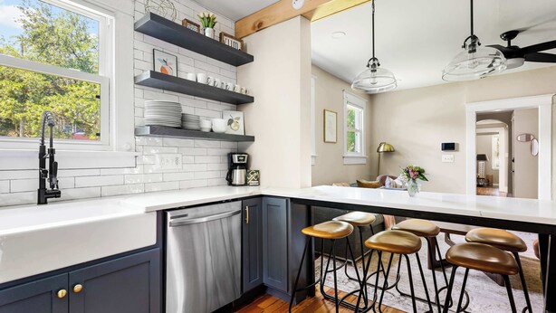A modern kitchen with a white countertop, dark cabinets, and a wooden floor. There are bar stools and a ceiling fan.