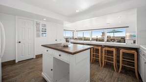 Una cocina moderna con una isla central, taburetes de bar y grandes ventanales con vistas a la playa.