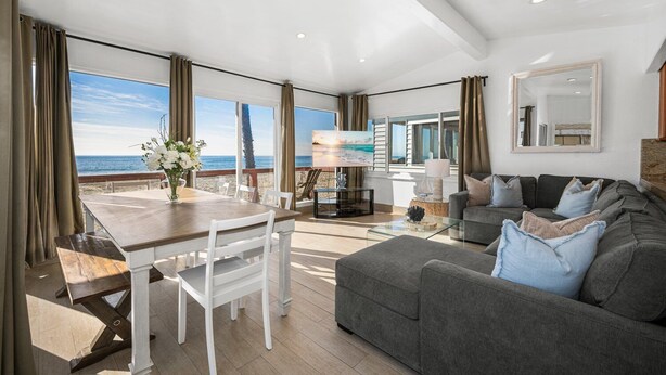 A spacious living area with a large window overlooking the ocean, a dining table with chairs, a gray sofa with cushions, and a mounted TV. - Oceanfront Home with Stunning Balcony Views (Newport Beach)