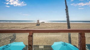 A beach view from a balcony with a lifeguard tower, palm trees, and a sandy beach. - Oceanfront Home with Stunning Balcony Views (Newport Beach)