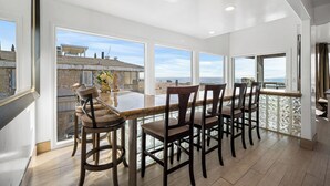 A spacious dining area with a large table and chairs, large windows offering a view of the beach and buildings, and a high ceiling with recessed lighting. - Oceanfront Home with Stunning Balcony Views (Newport Beach)