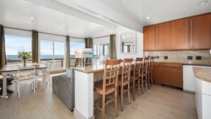 A modern kitchen with a central island, wooden chairs, a dining table, and large windows offering a view of the beach. - Oceanfront Home with Stunning Balcony Views (Newport Beach)