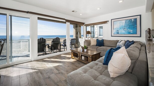 A spacious living room with a large window overlooking the beach, a comfortable sofa, and a wooden coffee table. - 5-Bedroom Oceanfront Escape with Private Balcony (Newport Beach)